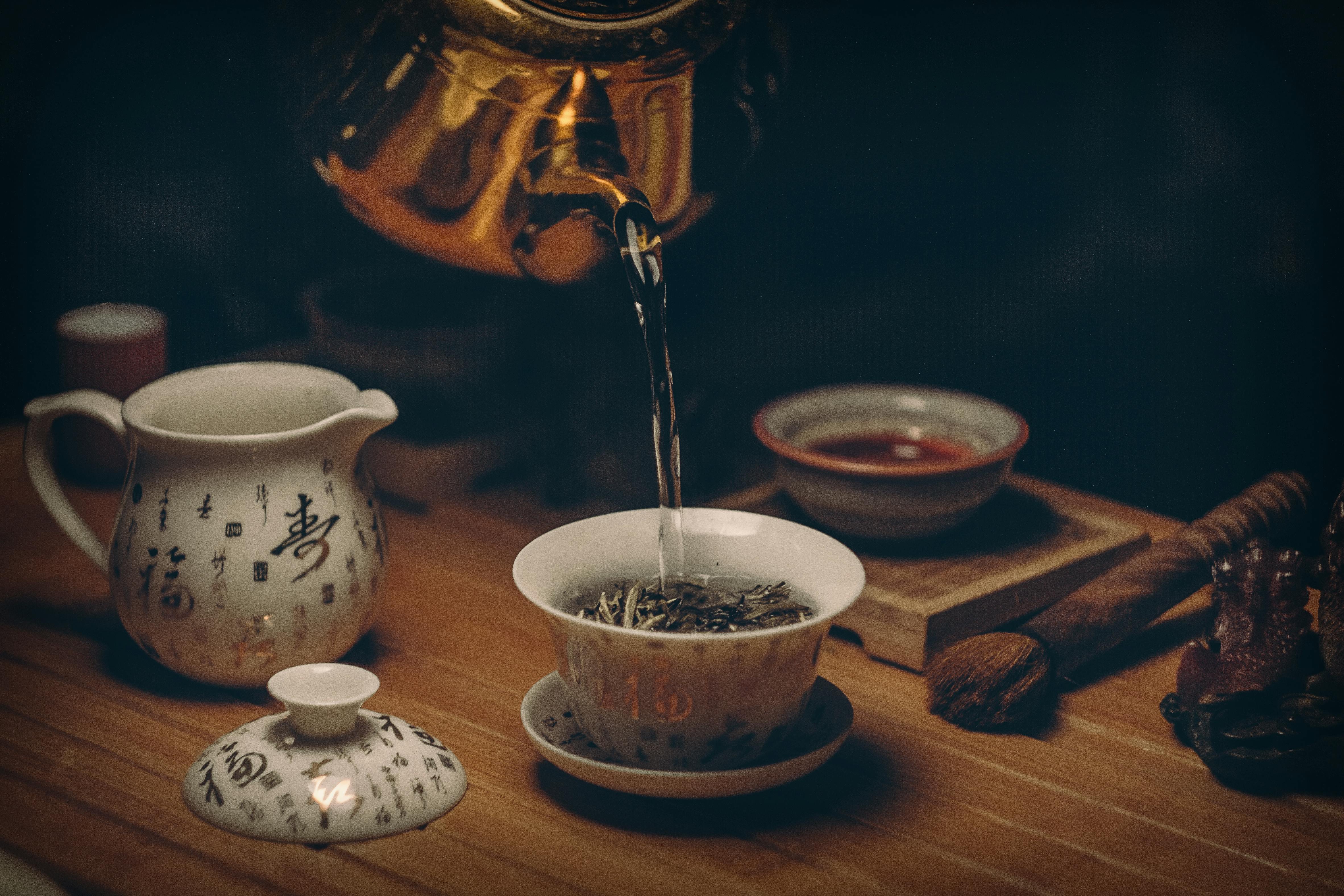 Golden teapot pouring hot water into a white ceramic bowl of loose-leaf tea on a wooden tea tray